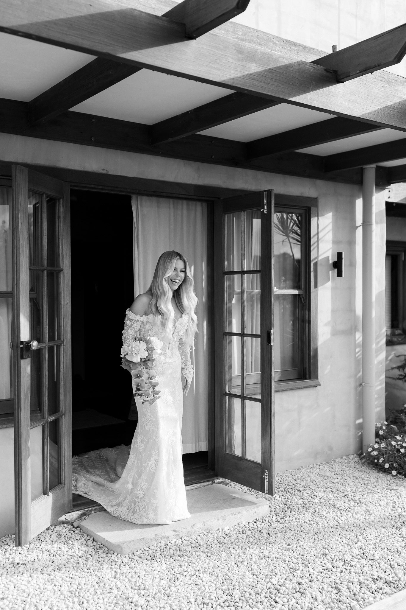 Black and white photo of a smiling bride in lace gown holding bouquet, standing in doorway of rustic venue.