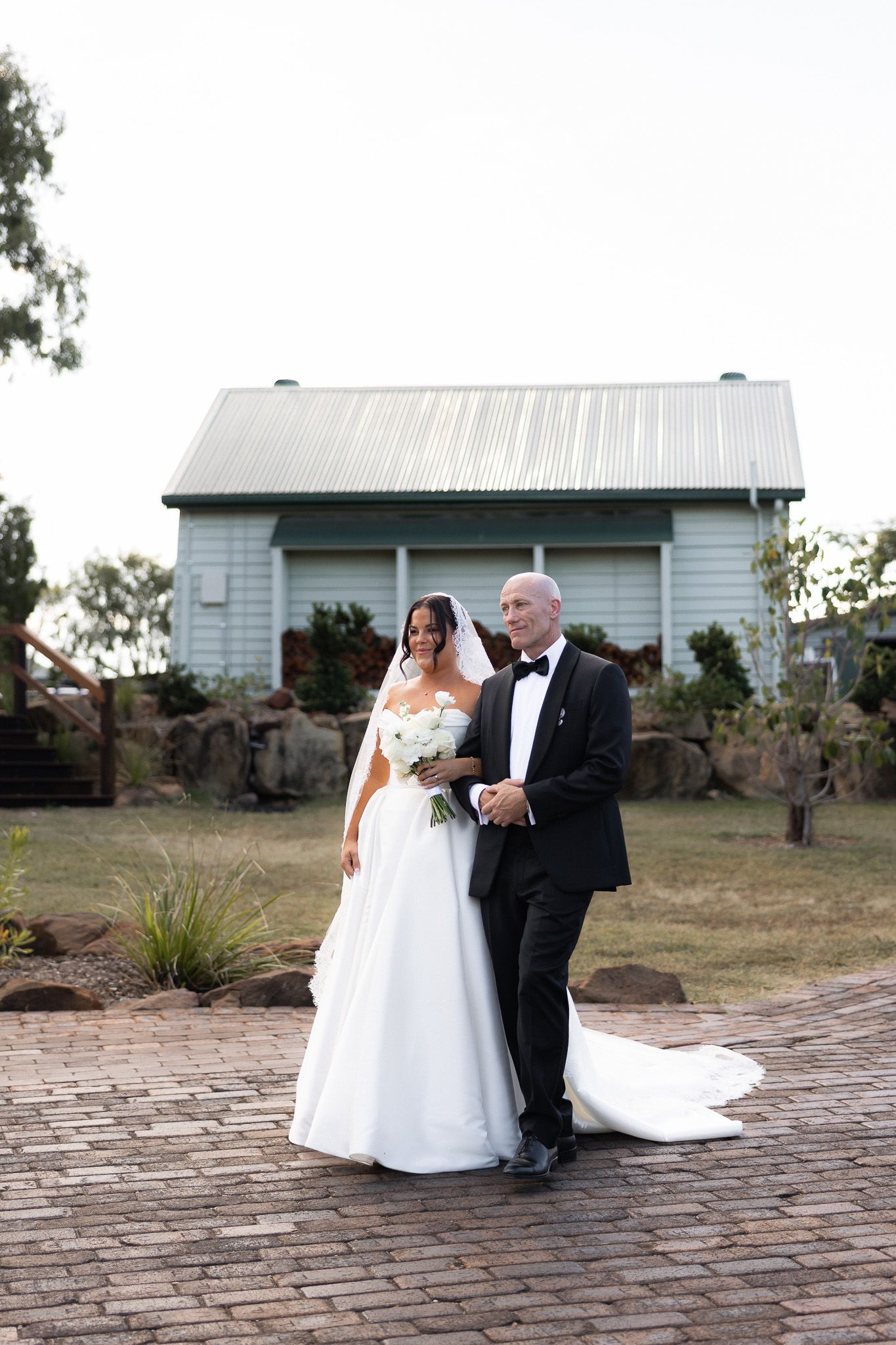 Bride in white gown and veil walking arm in arm with father toward wedding ceremony venue.