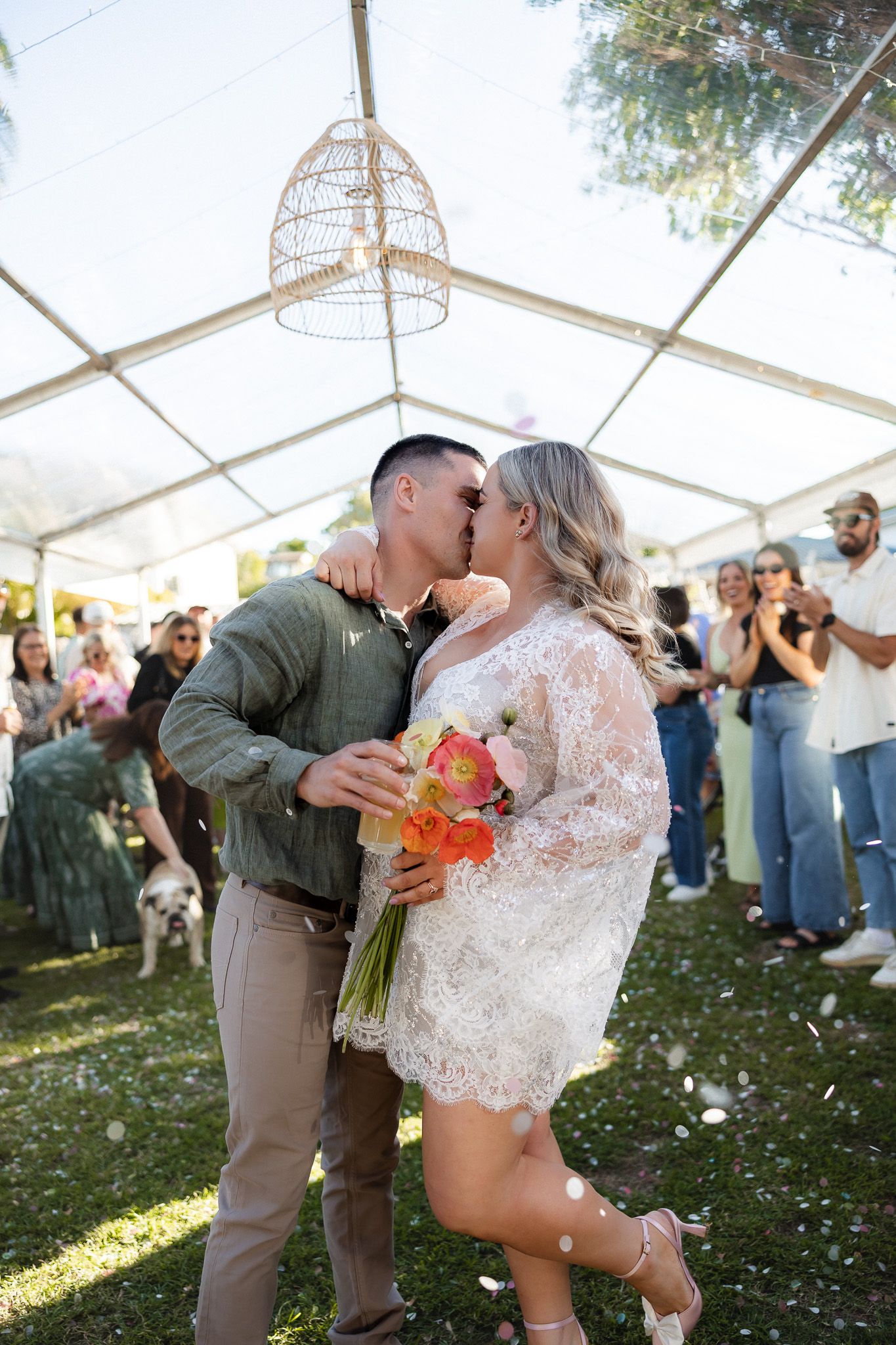 Bride and groom share a kiss under a tent at an outdoor wedding ceremony, with guests celebrating around them.