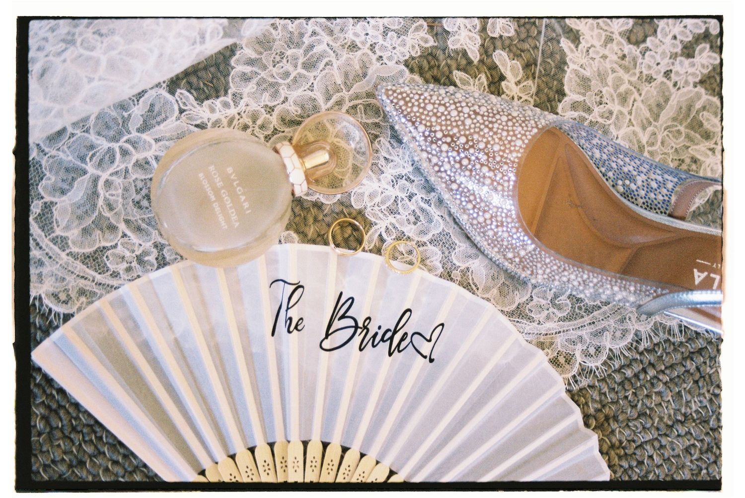 Bridal flat lay featuring a crystal heel, perfume bottle, lace fabric, gold rings, and a white The Bride fan.