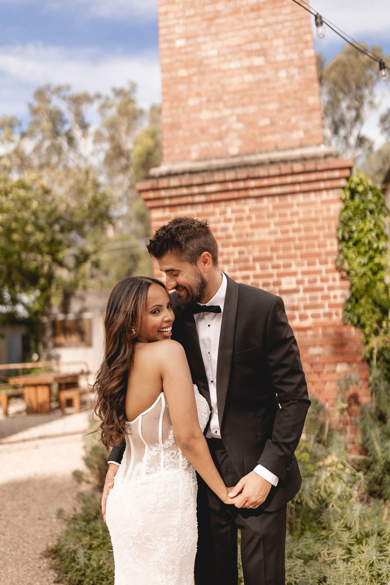 Bride in lace wedding gown and groom in black tuxedo embrace romantically outdoors near brick chimney.