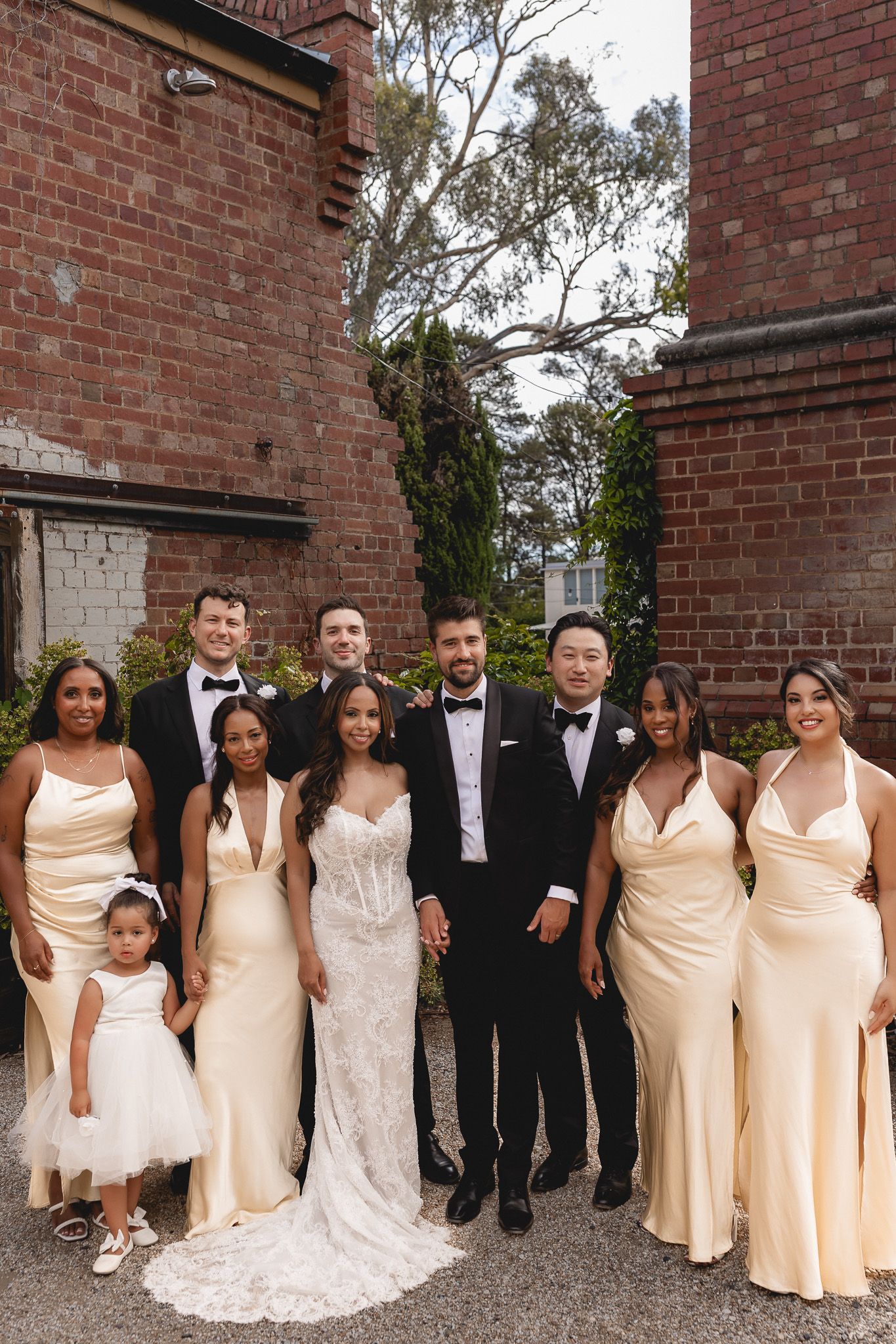 Wedding party poses in cream gowns and tuxedos before a brick wall with lush greenery.