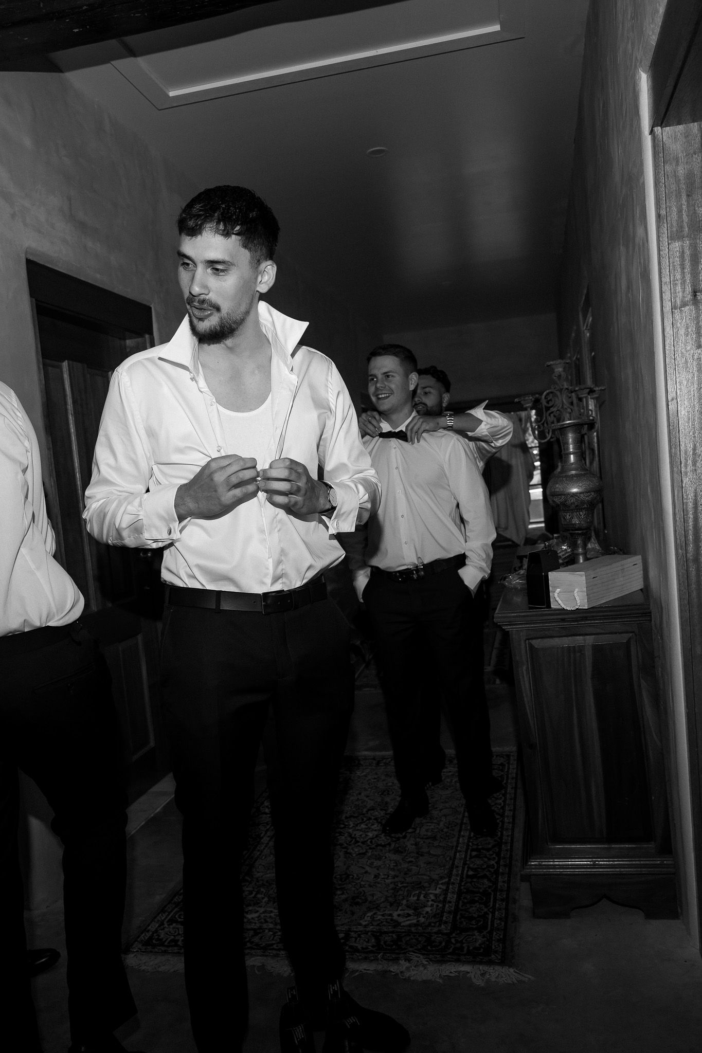 Black and white photo of groomsmen getting dressed in suits before wedding ceremony indoors.