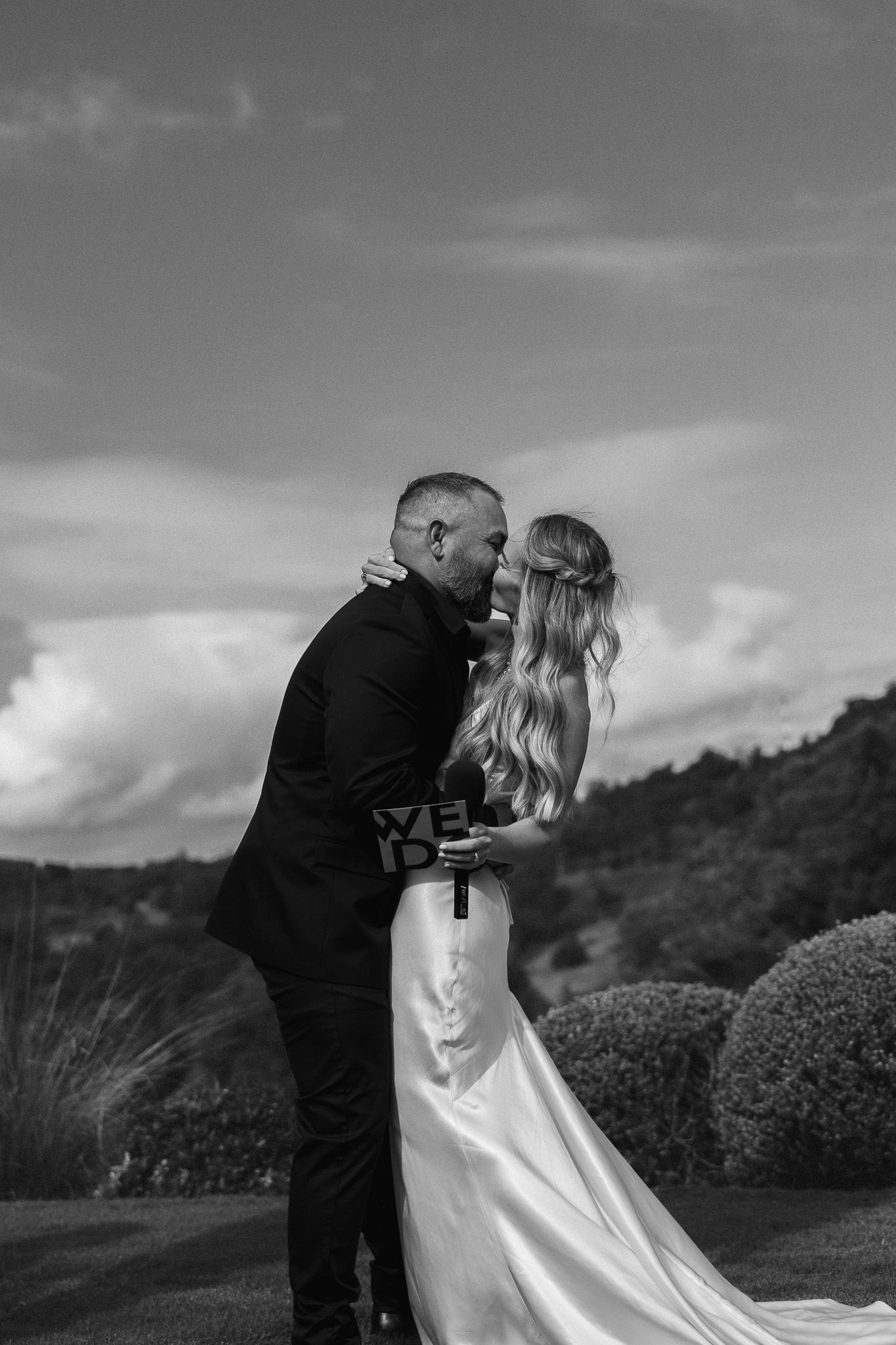 Black and white photo of a bride and groom sharing a romantic kiss outdoors on their wedding day.