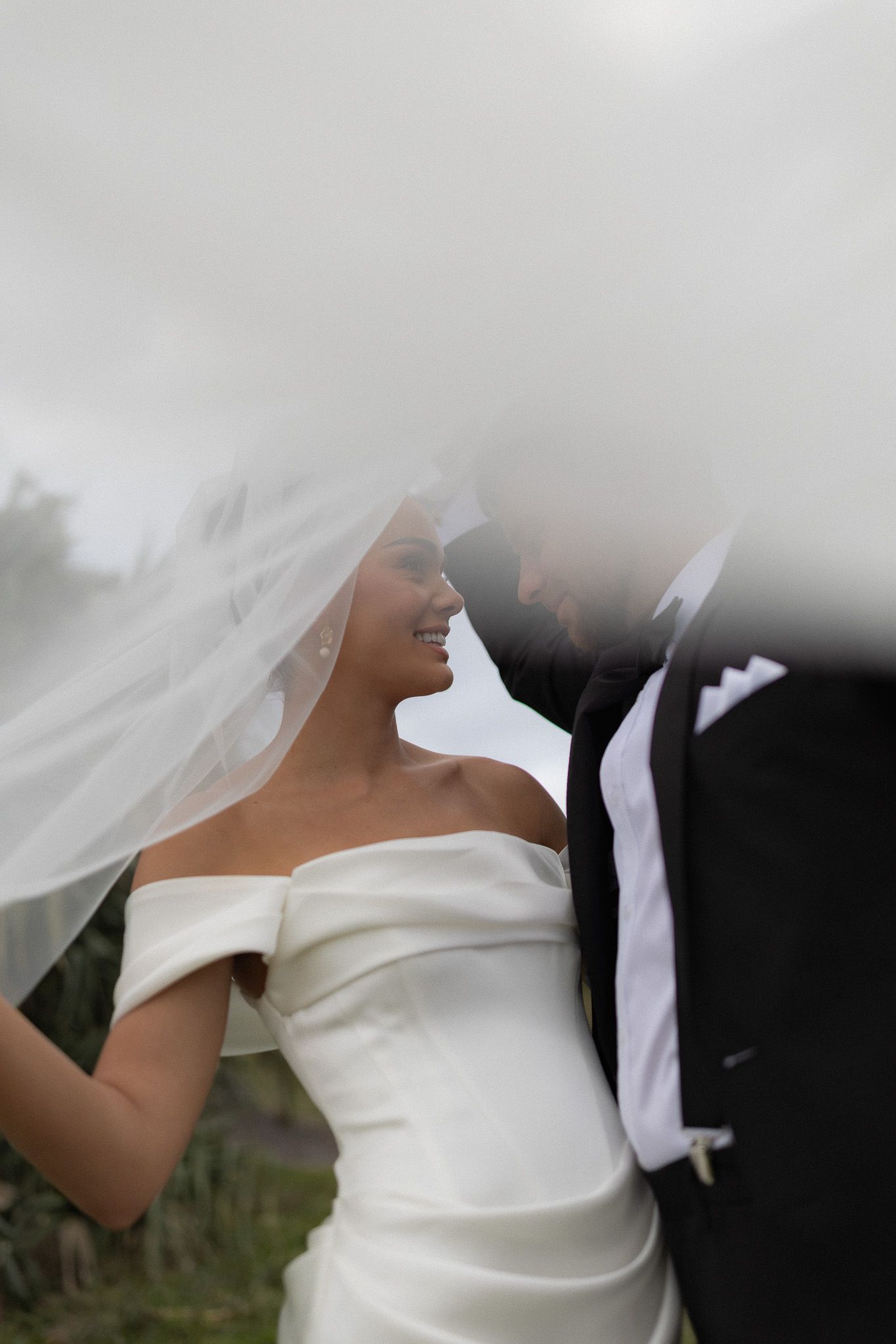 Bride smiles beneath flowing veil as groom leans in close during romantic outdoor wedding portrait.
