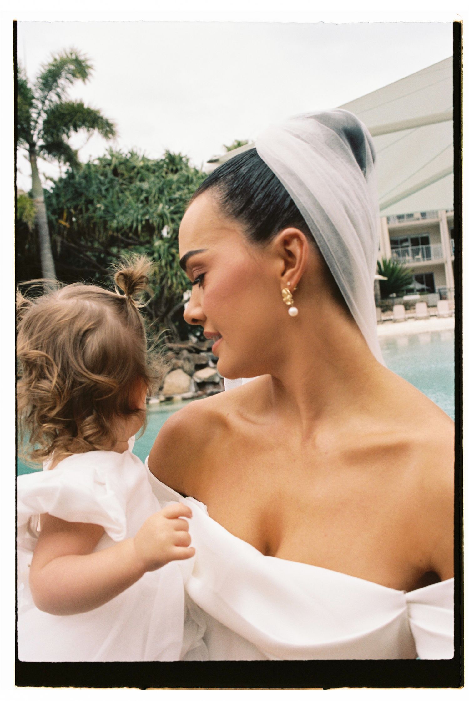 Bride in off-shoulder gown and veil holds young child in white dress near a pool.