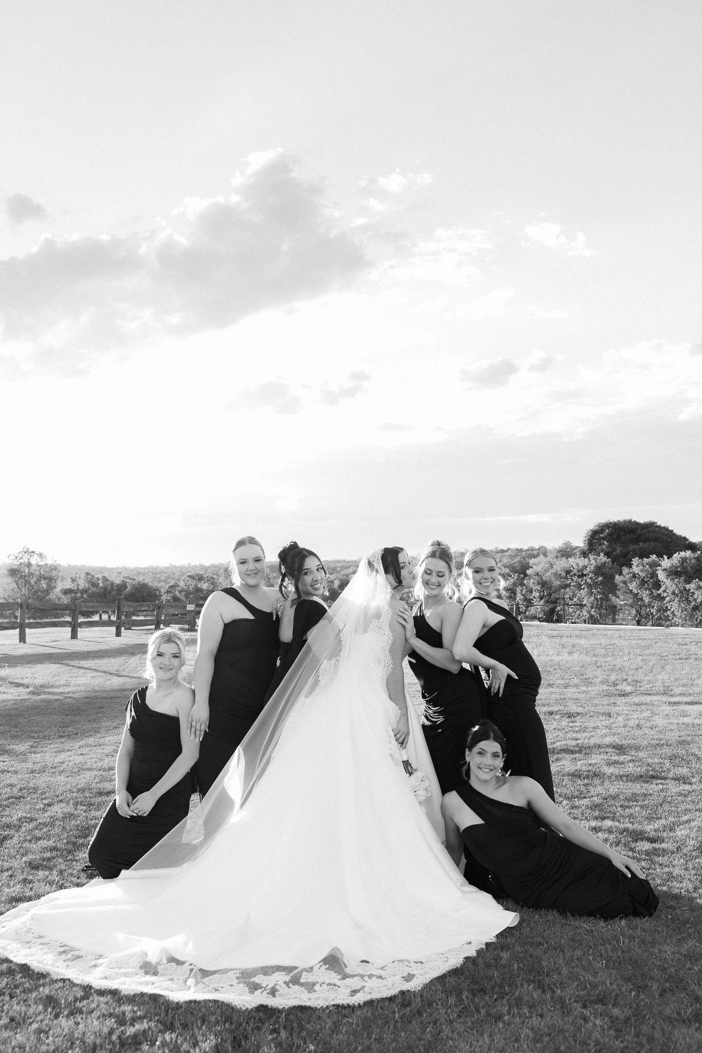 Black and white photo of a bride in a white gown and veil surrounded by bridesmaids in black dresses on a grassy lawn.