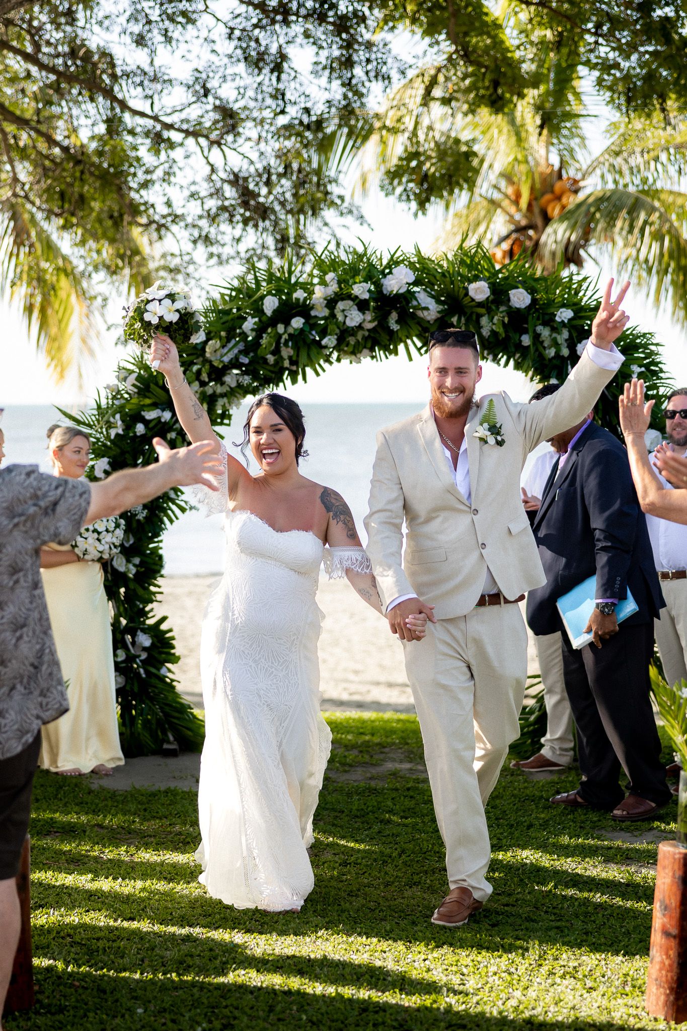 Newlyweds celebrate joyfully under a floral arch at a beachside wedding ceremony with guests cheering behind them.