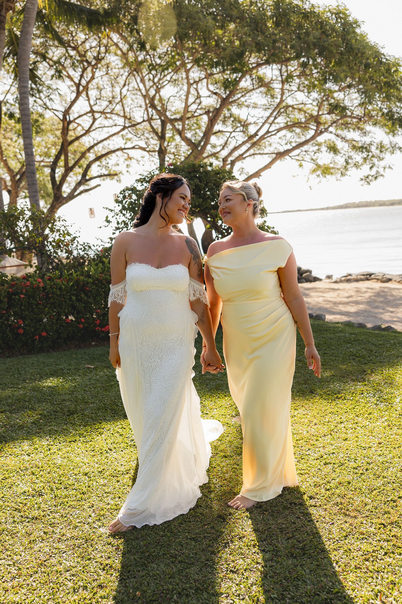Bride in white lace off-shoulder gown walking with bridesmaid in yellow dress near tropical beach at sunset.