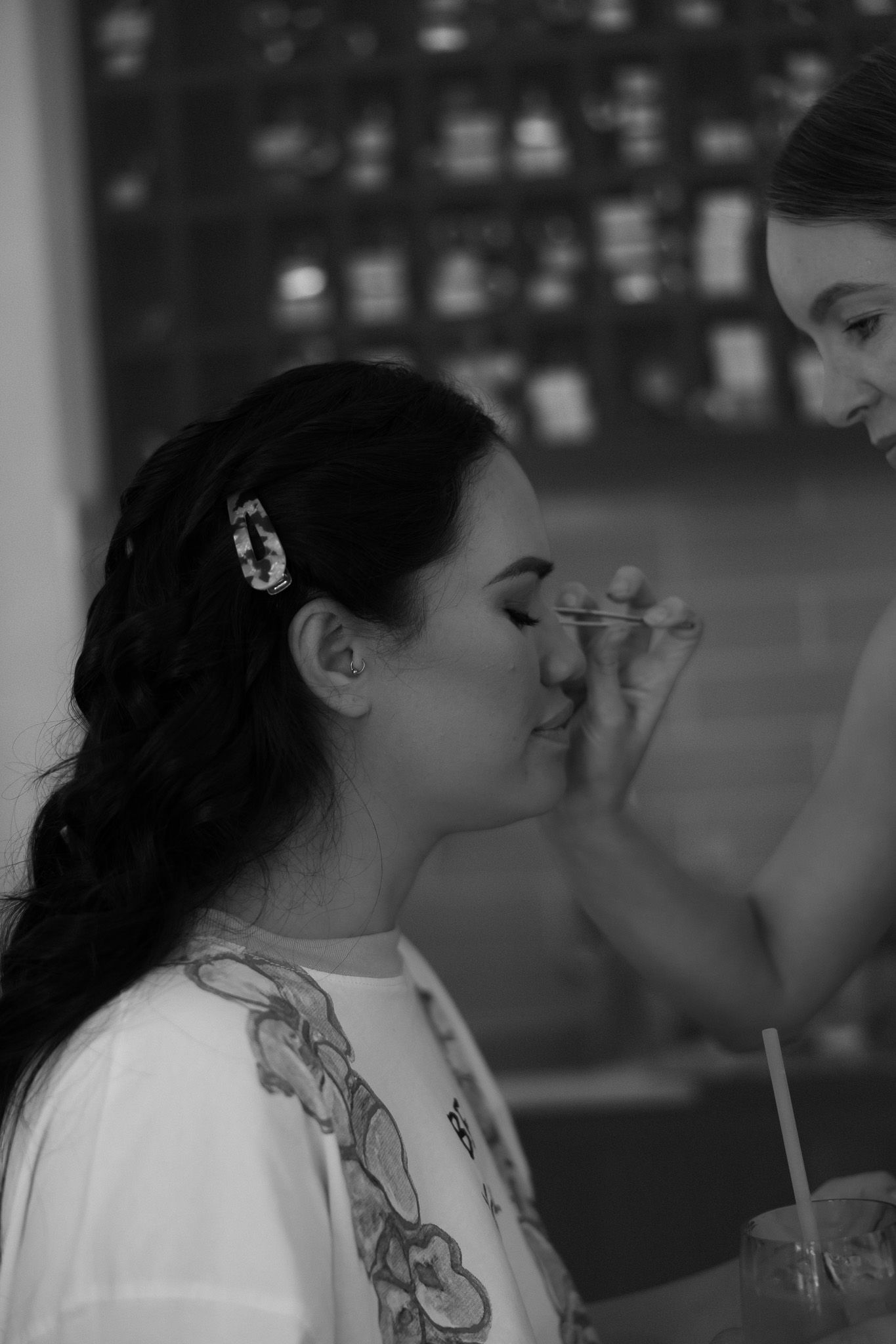 Black and white photo of a woman getting makeup applied, wearing a white top with hair clip.