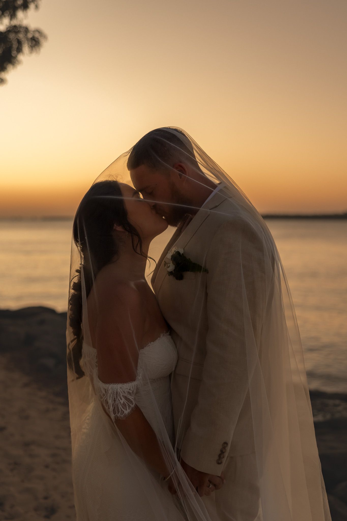 Bride and groom share romantic kiss under veil at golden sunset on beach.