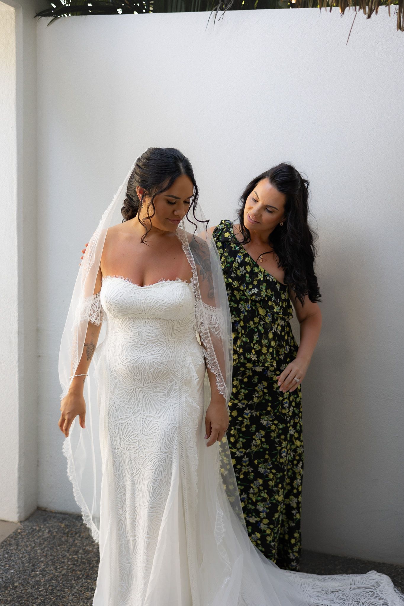 Bride in lace wedding gown and veil being helped by bridesmaid in floral dress against white wall.