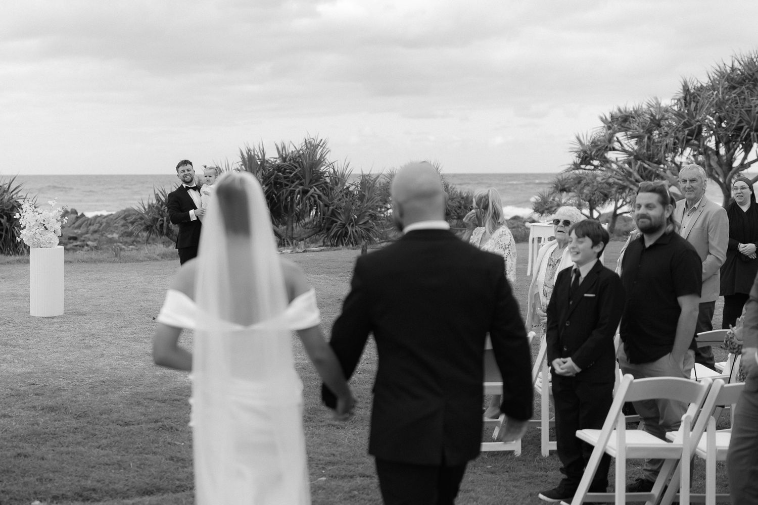 Black and white photo of bride walking down aisle toward waiting groom outdoors.