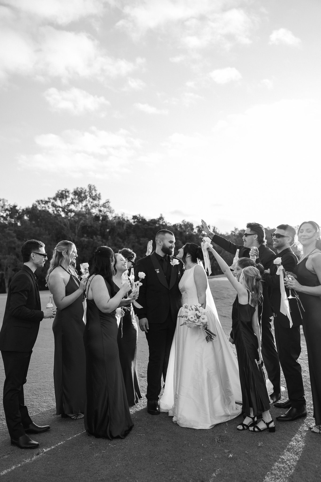 Black and white wedding party photo with bride and groom surrounded by bridesmaids and groomsmen raising glasses outdoors.