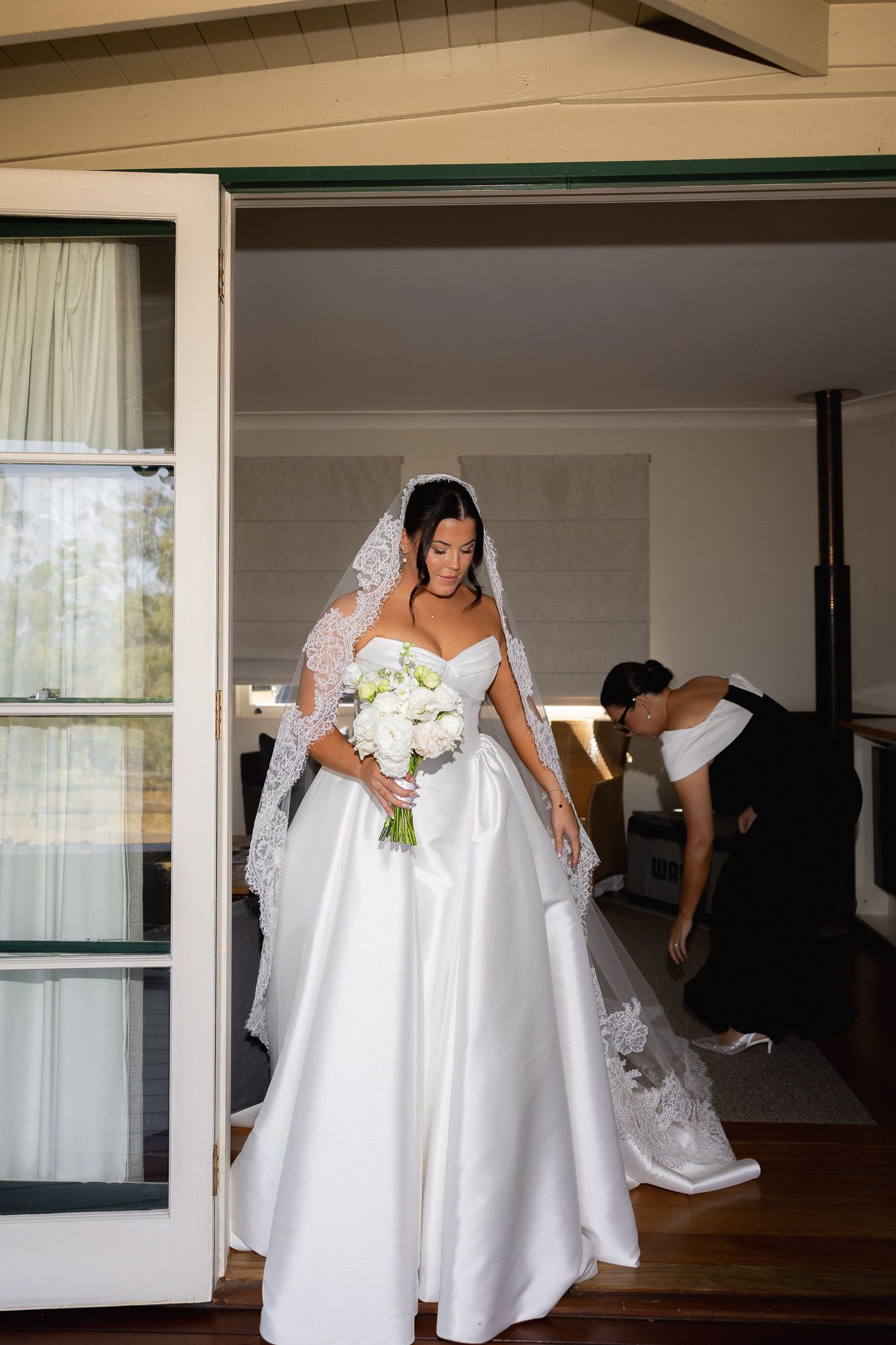 Elegant bride in strapless ball gown with lace veil holds white bouquet as assistant adjusts train.