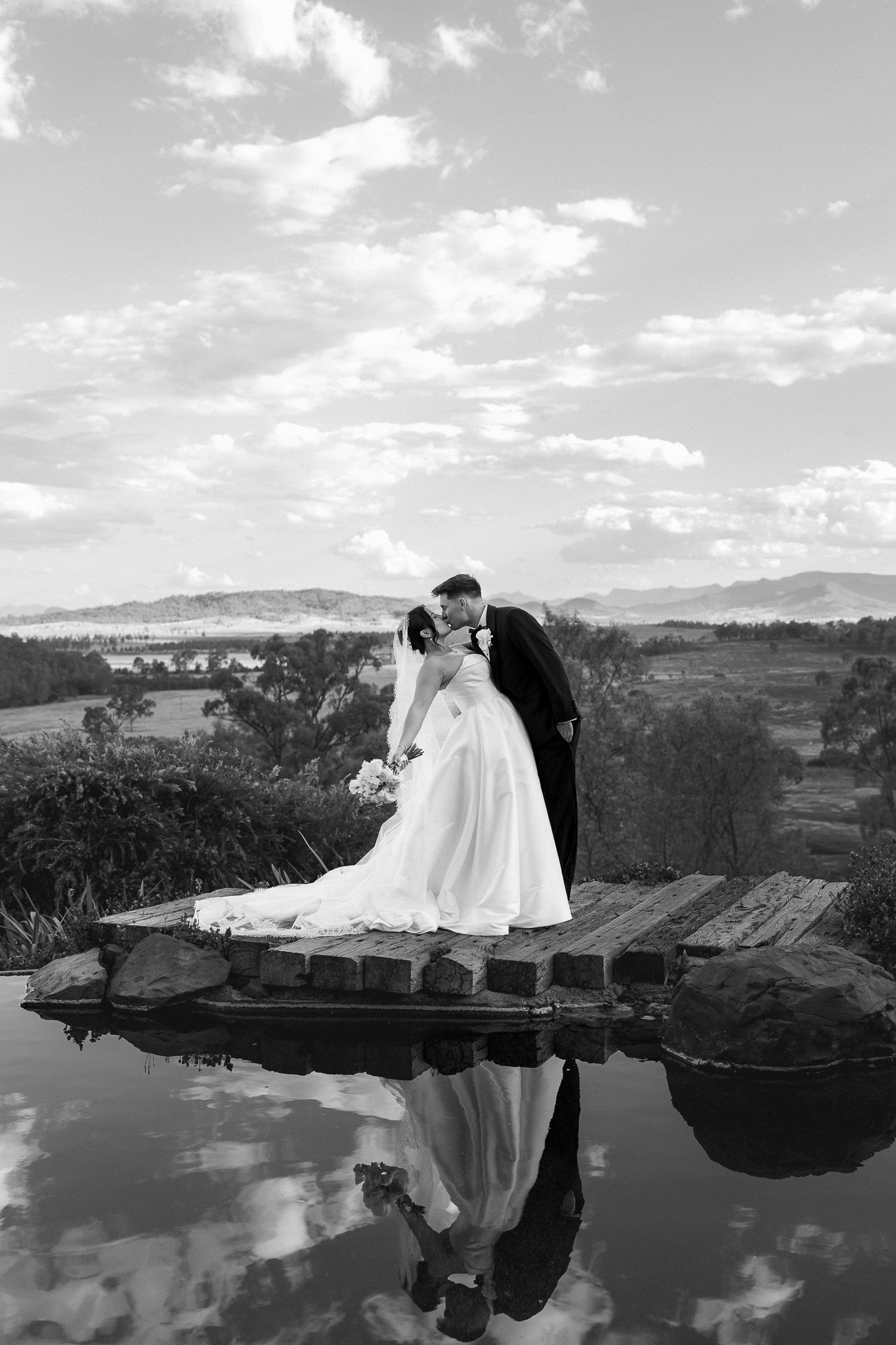 Black and white photo of bride and groom kissing on a dock with their reflection mirrored in still water below.