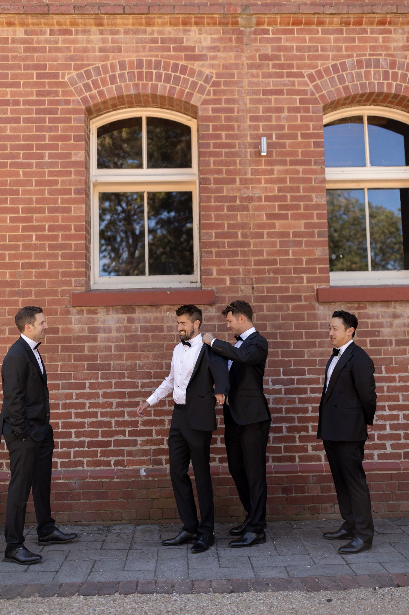 Four men in formal black suits stand against a red brick building wall, with two groomsmen adjusting the groom's attire.