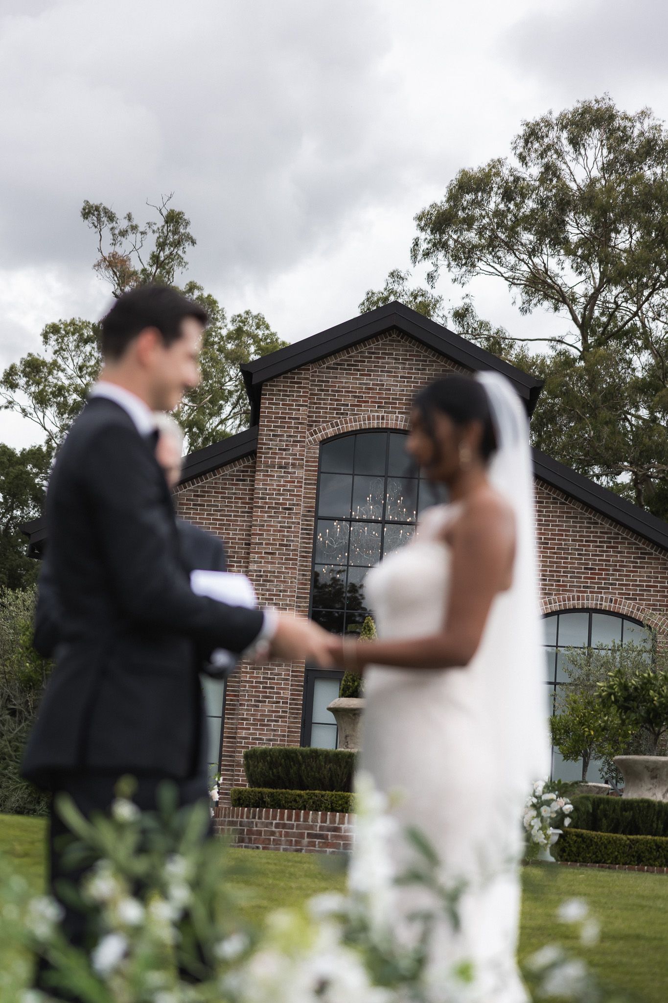 Bride and groom exchanging vows outdoors in front of elegant brick wedding venue with lush gardens.