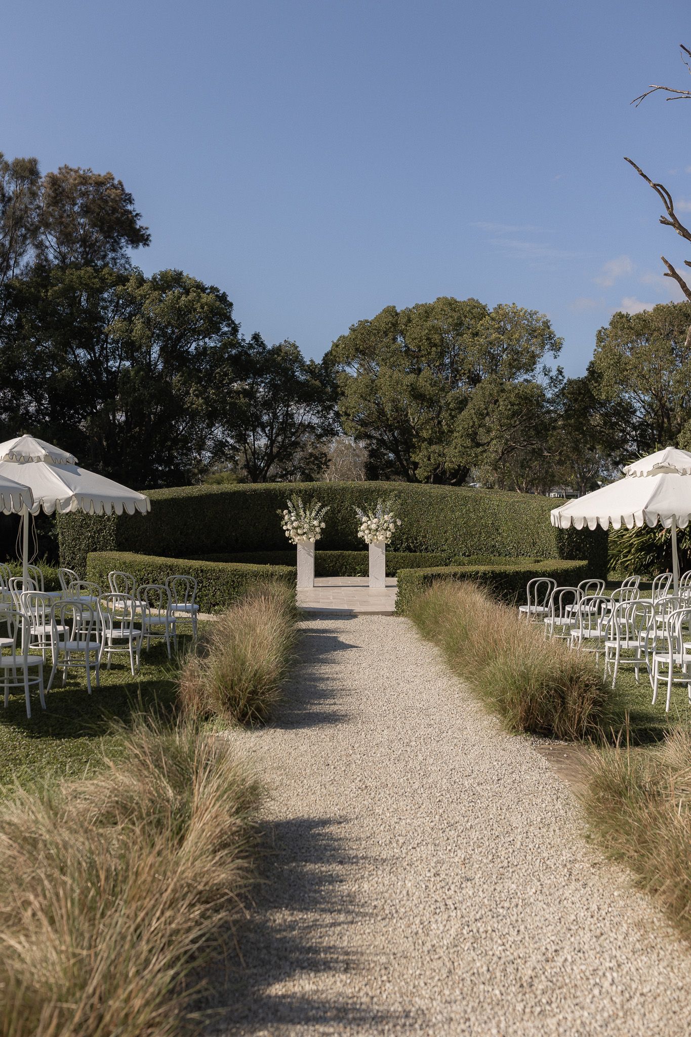 Elegant outdoor wedding ceremony setup with white chairs, gravel aisle, floral arch, trimmed hedges, and striped umbrellas under blue sky.