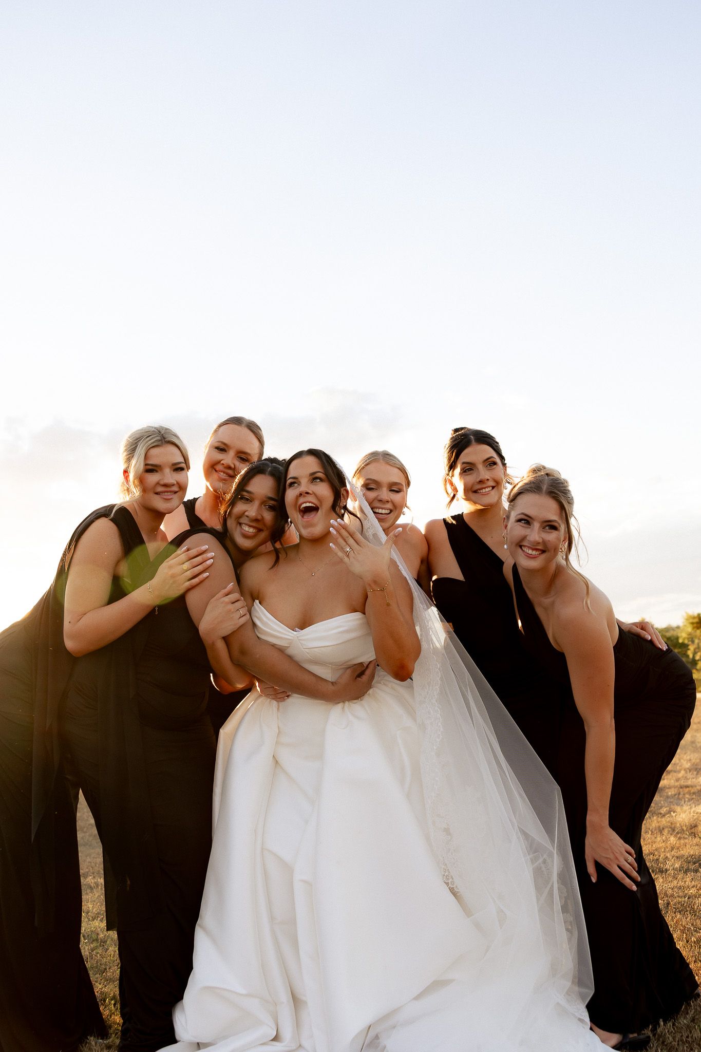 Laughing bride in white gown poses with six bridesmaids in black dresses during golden hour outdoors.