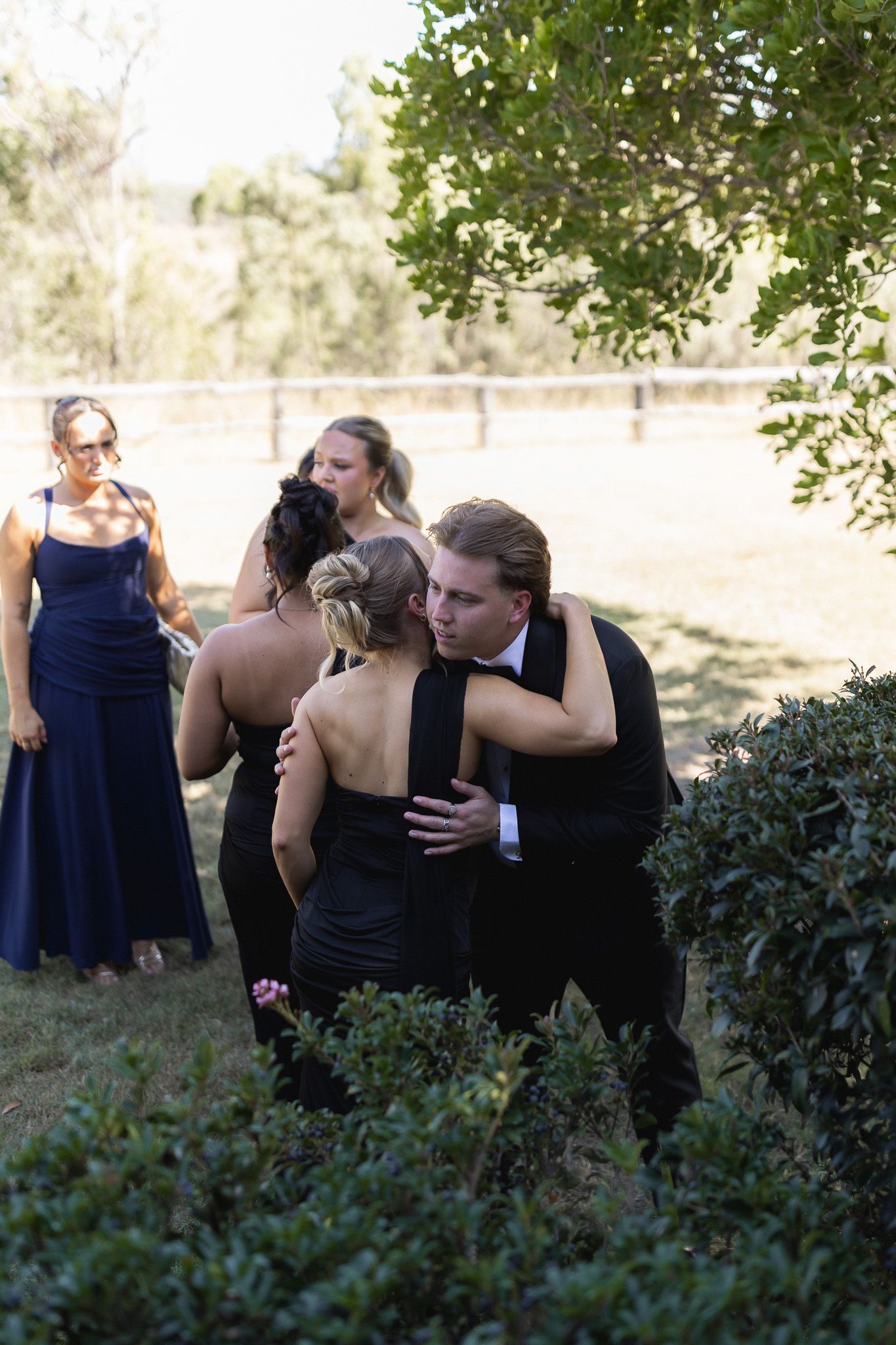 Man in tuxedo embracing woman in black dress at outdoor wedding reception with bridesmaids in navy blue gowns.