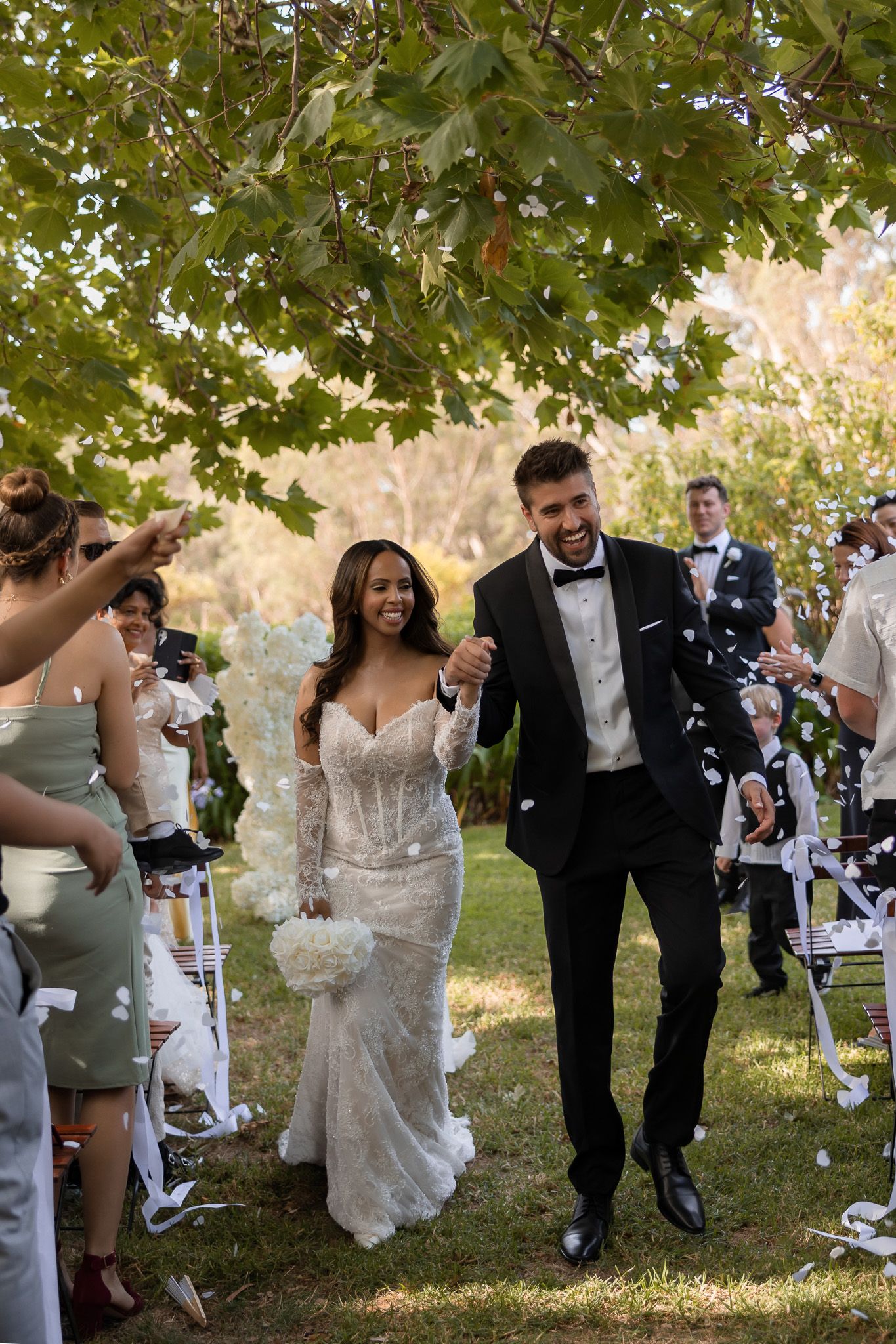 Bride and groom walk down outdoor aisle as guests toss white confetti during joyful wedding ceremony.