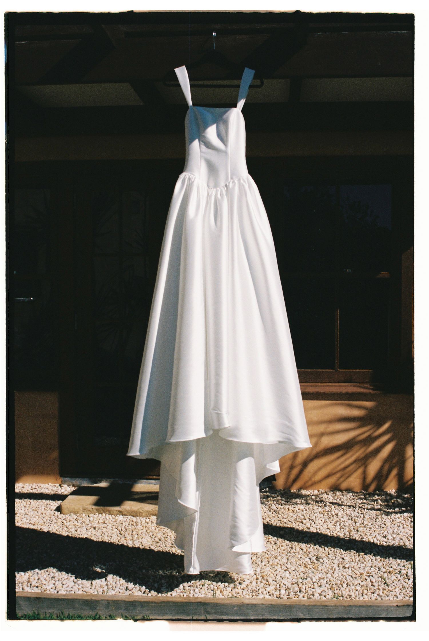 Elegant white bridal gown with high-low skirt hanging on a hanger outdoors in natural sunlight.