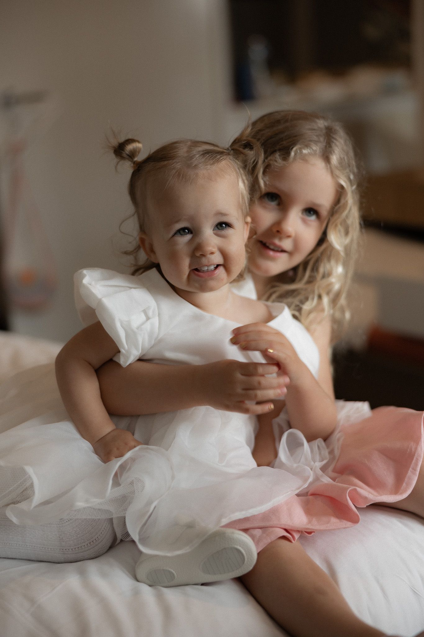 Two young girls in white dresses smile and hug on a bed.