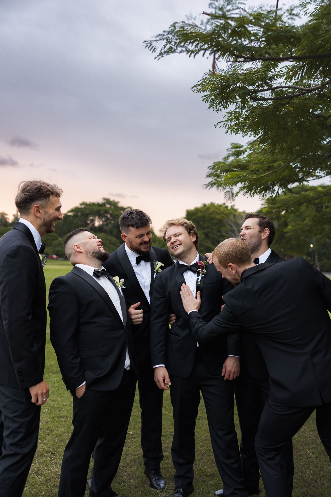 Six groomsmen in black tuxedos laughing together outdoors at dusk, with lush green trees in the background.