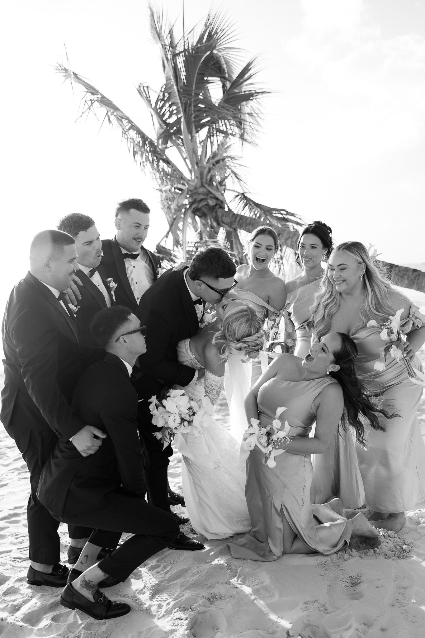 Black and white beach wedding party photo with bride, groom, groomsmen, and bridesmaids posing by a palm tree.
