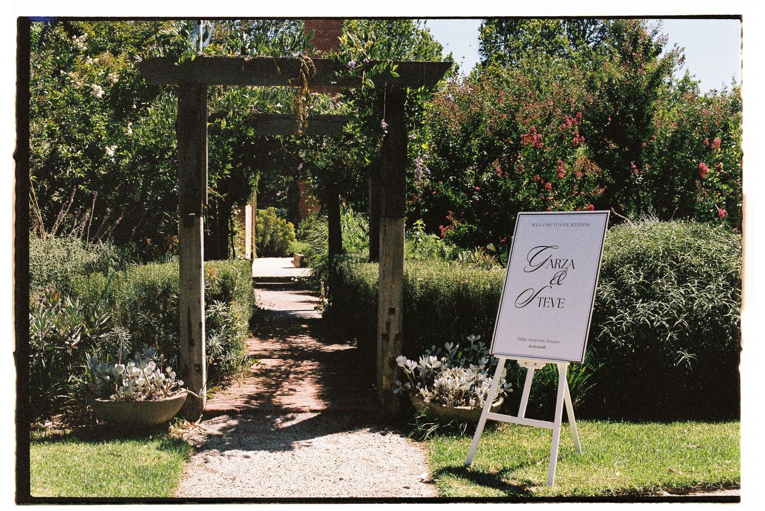 Garden wedding venue entrance with wooden pergola, lush greenery, and elegant welcome sign on easel.