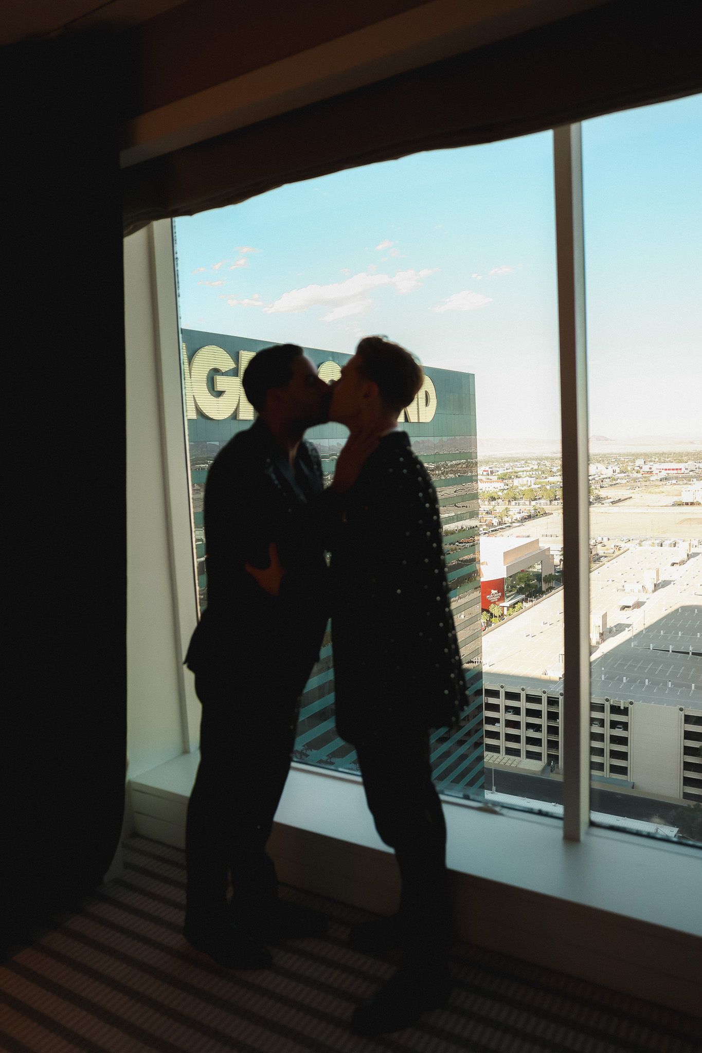 Two people share a romantic kiss silhouetted against a high-rise Las Vegas hotel window overlooking the city skyline.