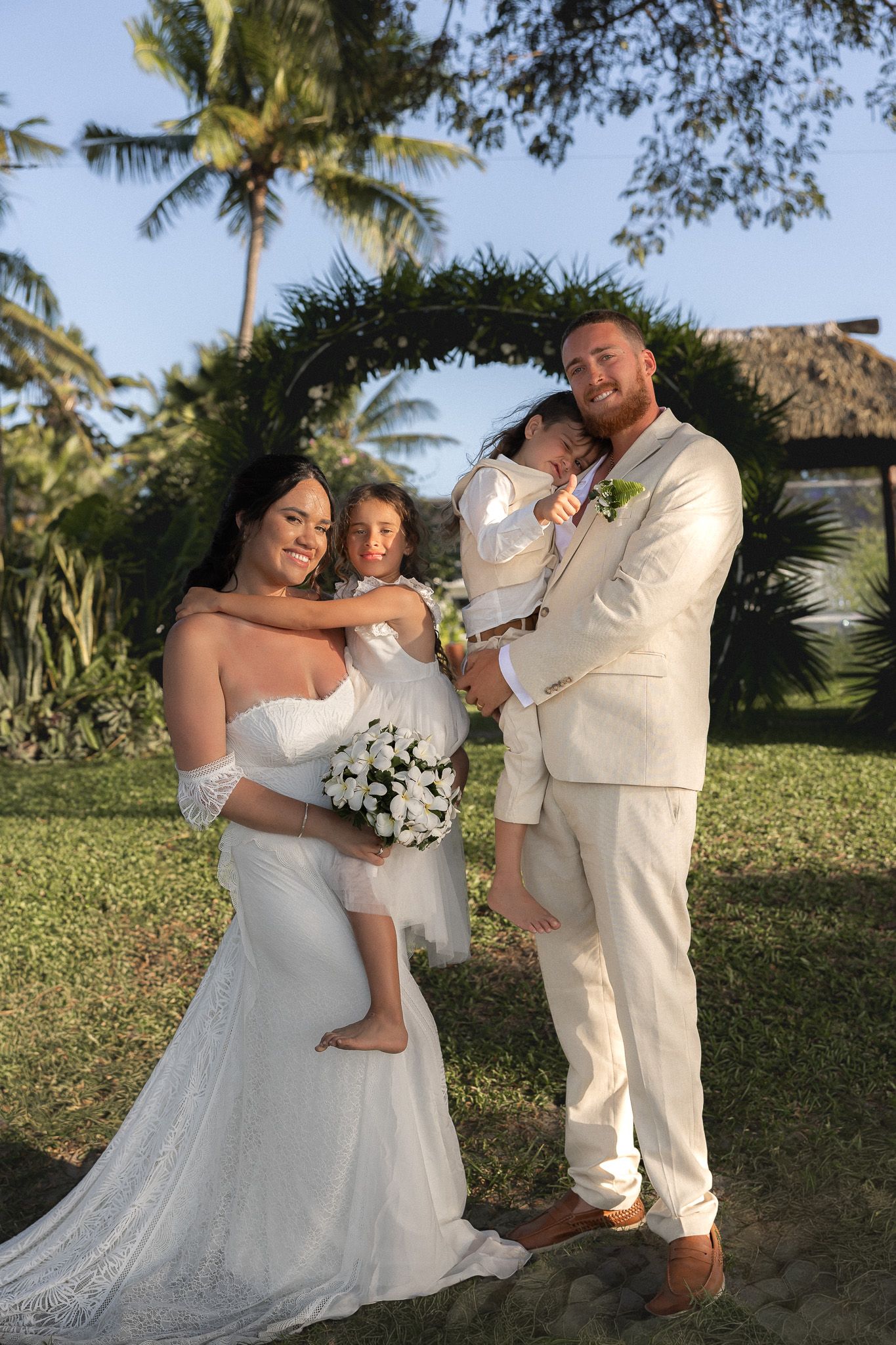 Bride and groom in cream suit hold two young children under floral arch at tropical wedding.