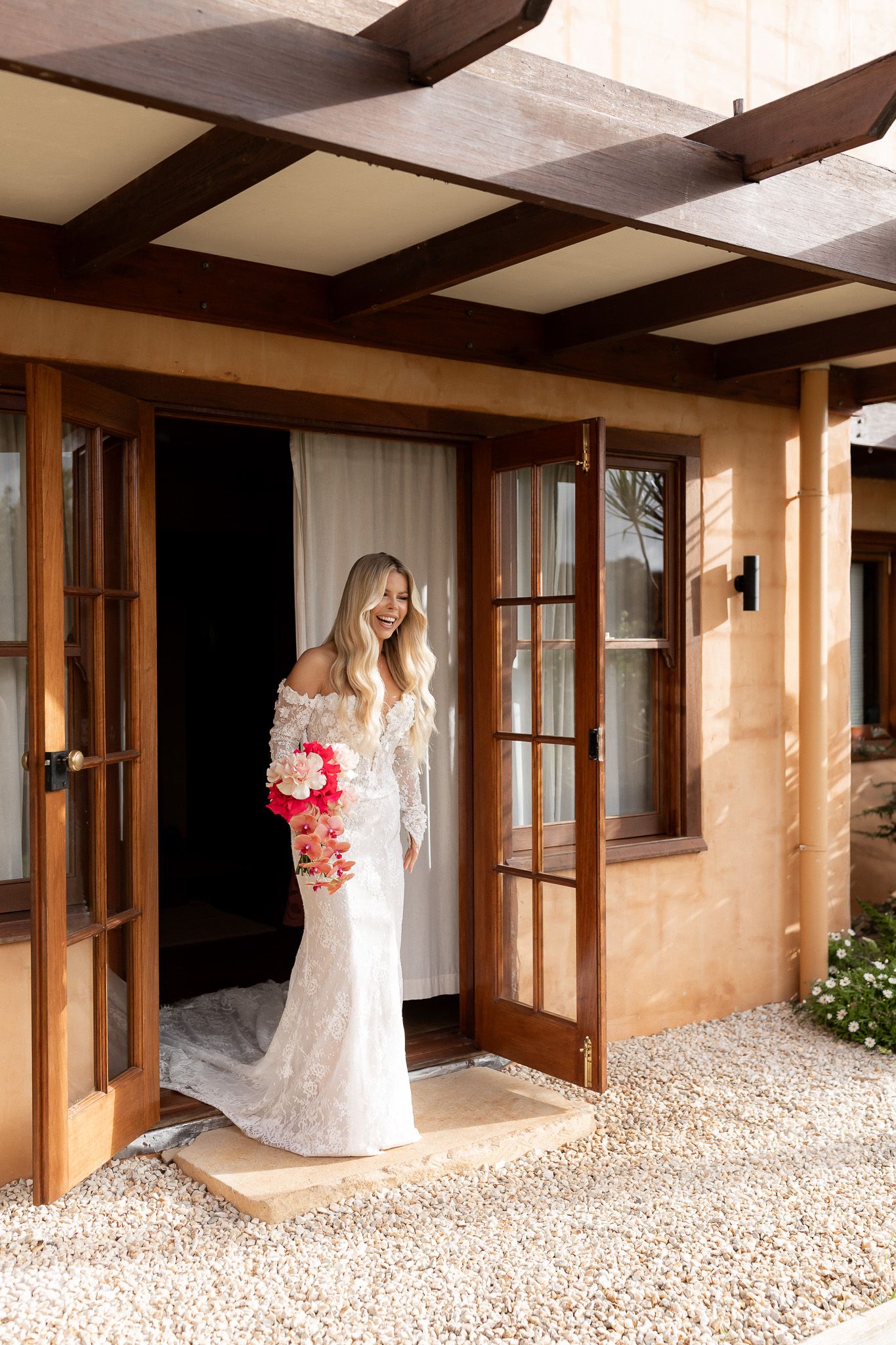 Bride in lace off-shoulder gown holding pink bouquet, standing in doorway of rustic adobe venue.