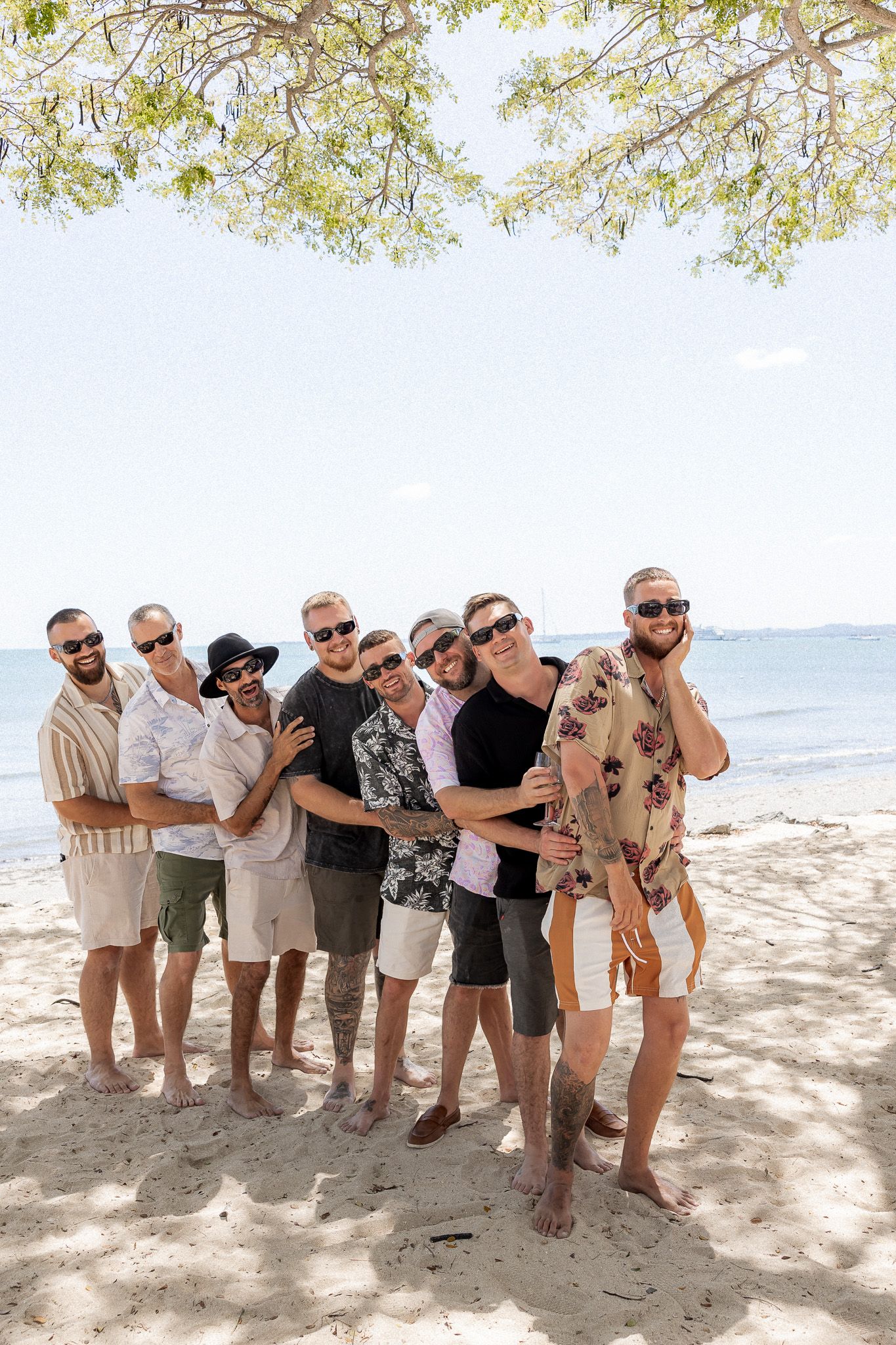Group of men posing together on a sandy beach, wearing sunglasses and casual summer outfits with ocean in background.