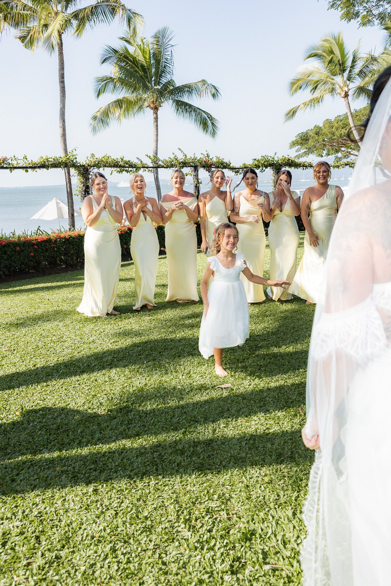 Bridesmaids in cream dresses clapping as flower girl in white runs toward the bride outdoors.