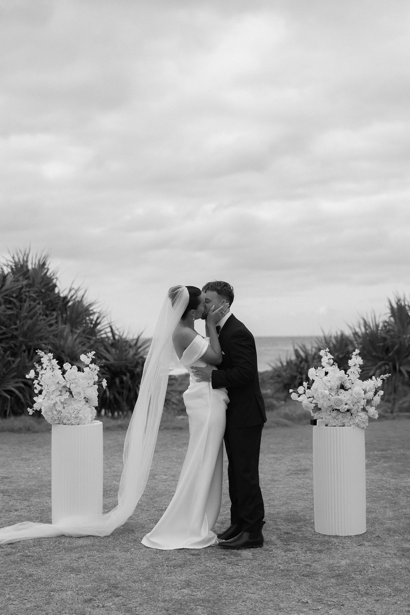 Black and white photo of bride and groom sharing a kiss at outdoor ceremony with floral pedestals.