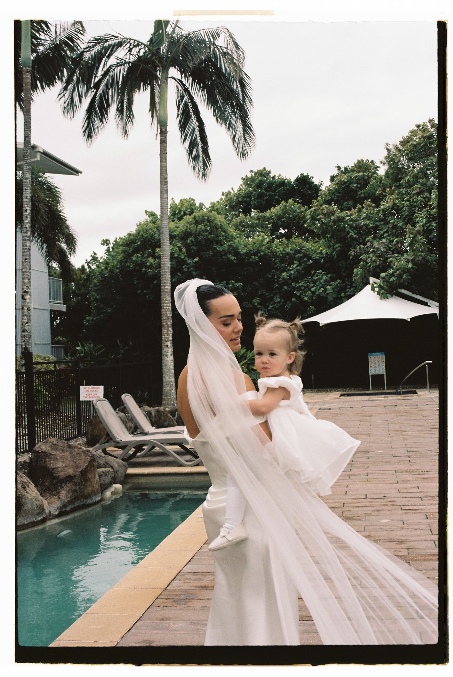 Bride in white gown and veil holds a toddler in white dress beside a tropical pool.