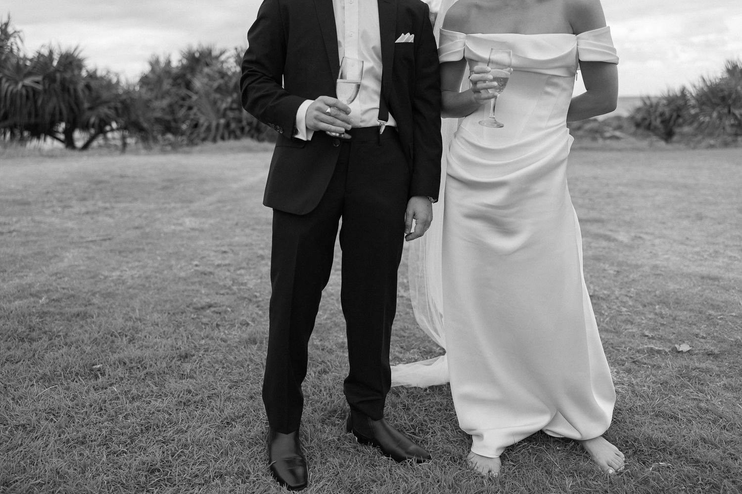 Black and white photo of bride in off-shoulder gown and groom in black suit holding champagne glasses outdoors.
