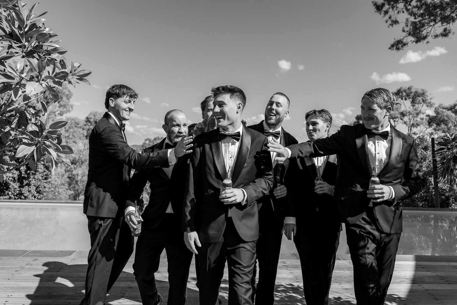 Groom and groomsmen in black tuxedos laughing and celebrating outdoors on a sunny day.