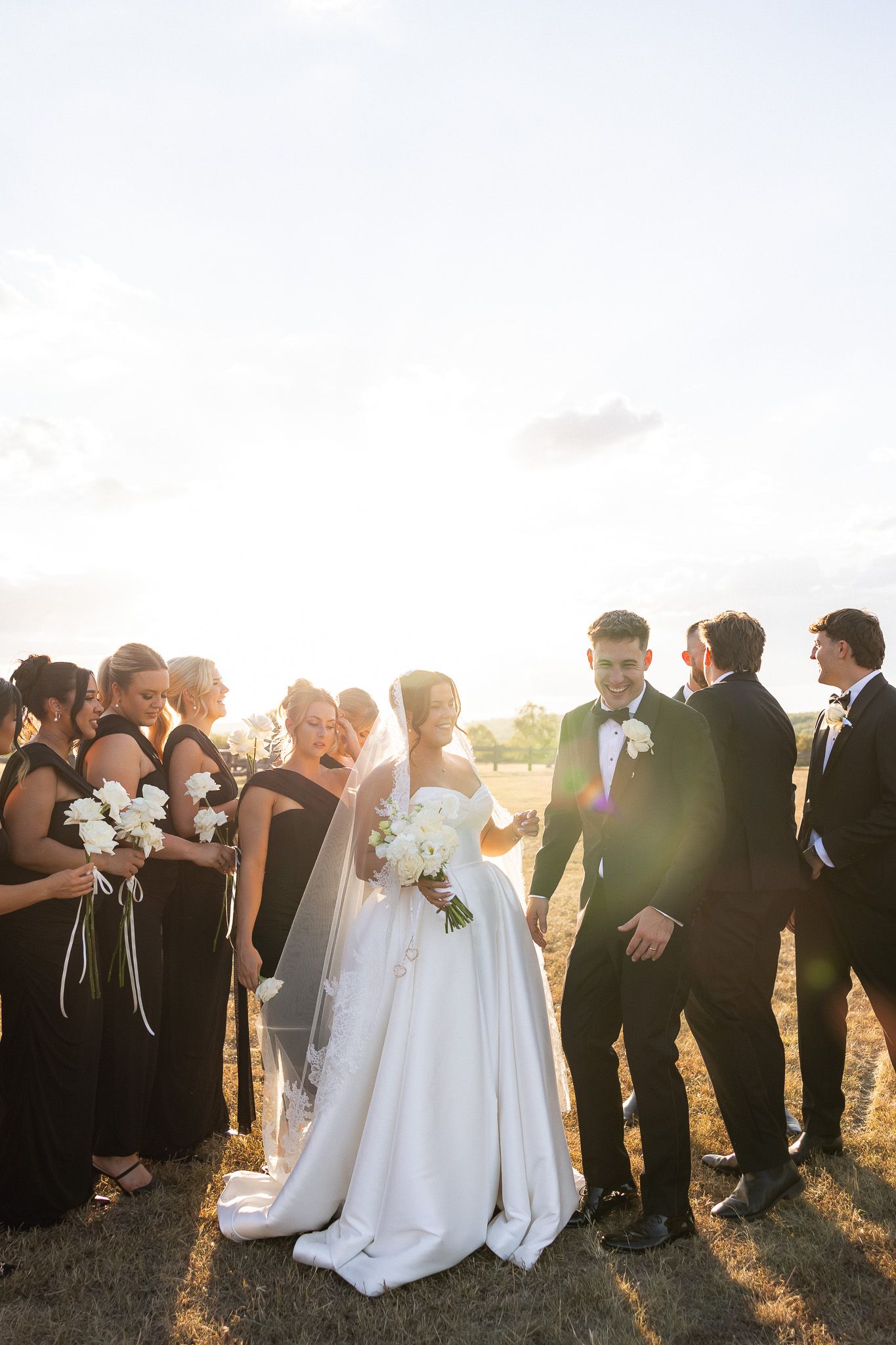 Bride and groom stand with bridesmaids in black dresses and groomsmen in tuxedos outdoors at golden hour.