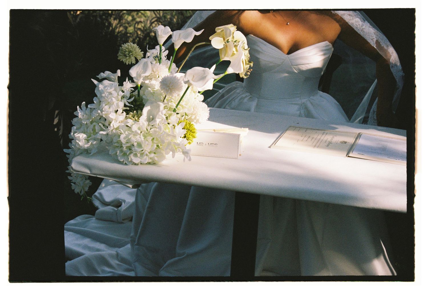 Bride in white strapless gown holding elegant white floral bouquet with orchids and daisies at outdoor wedding ceremony.