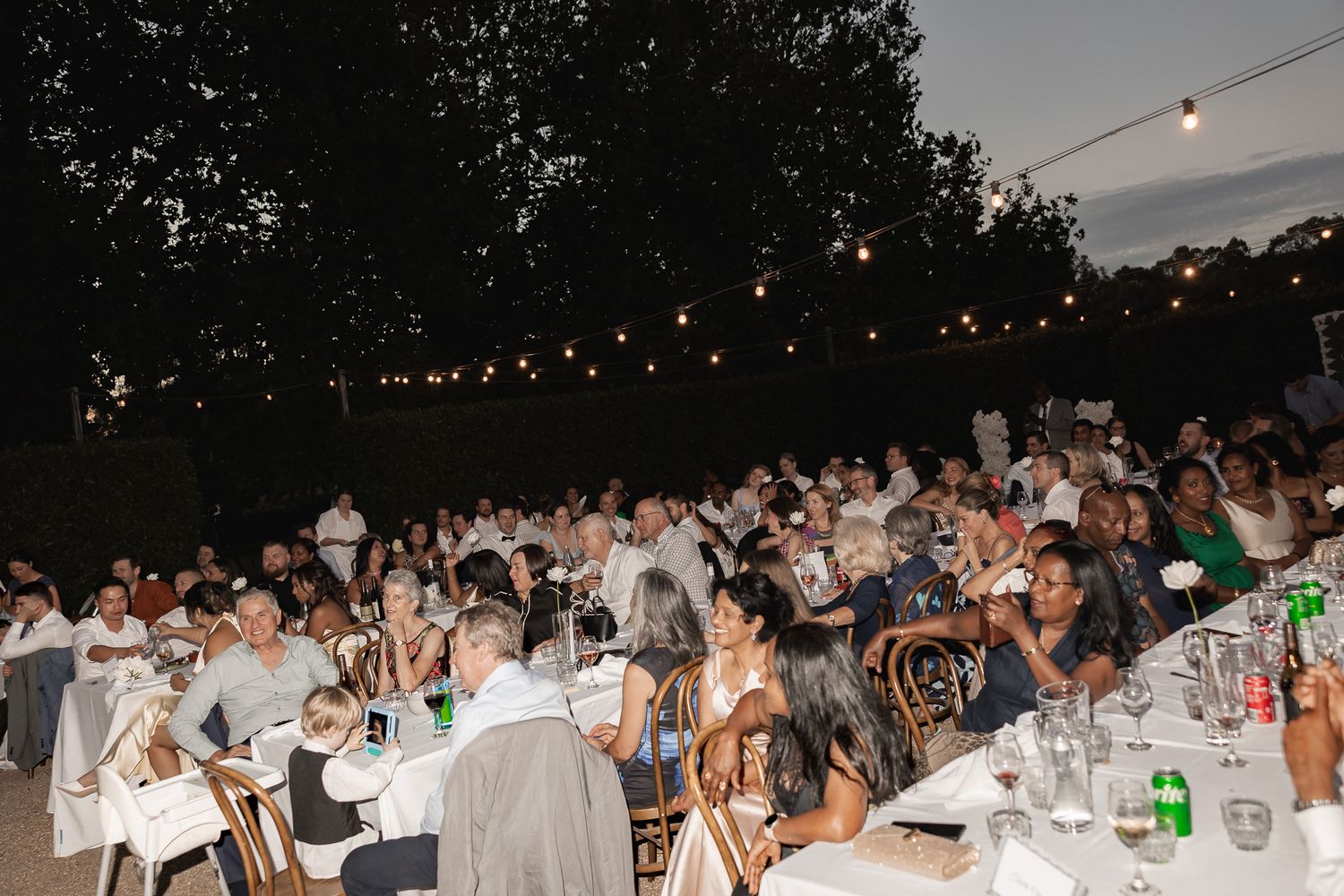 Large outdoor evening wedding reception with guests seated at white tables under string lights.