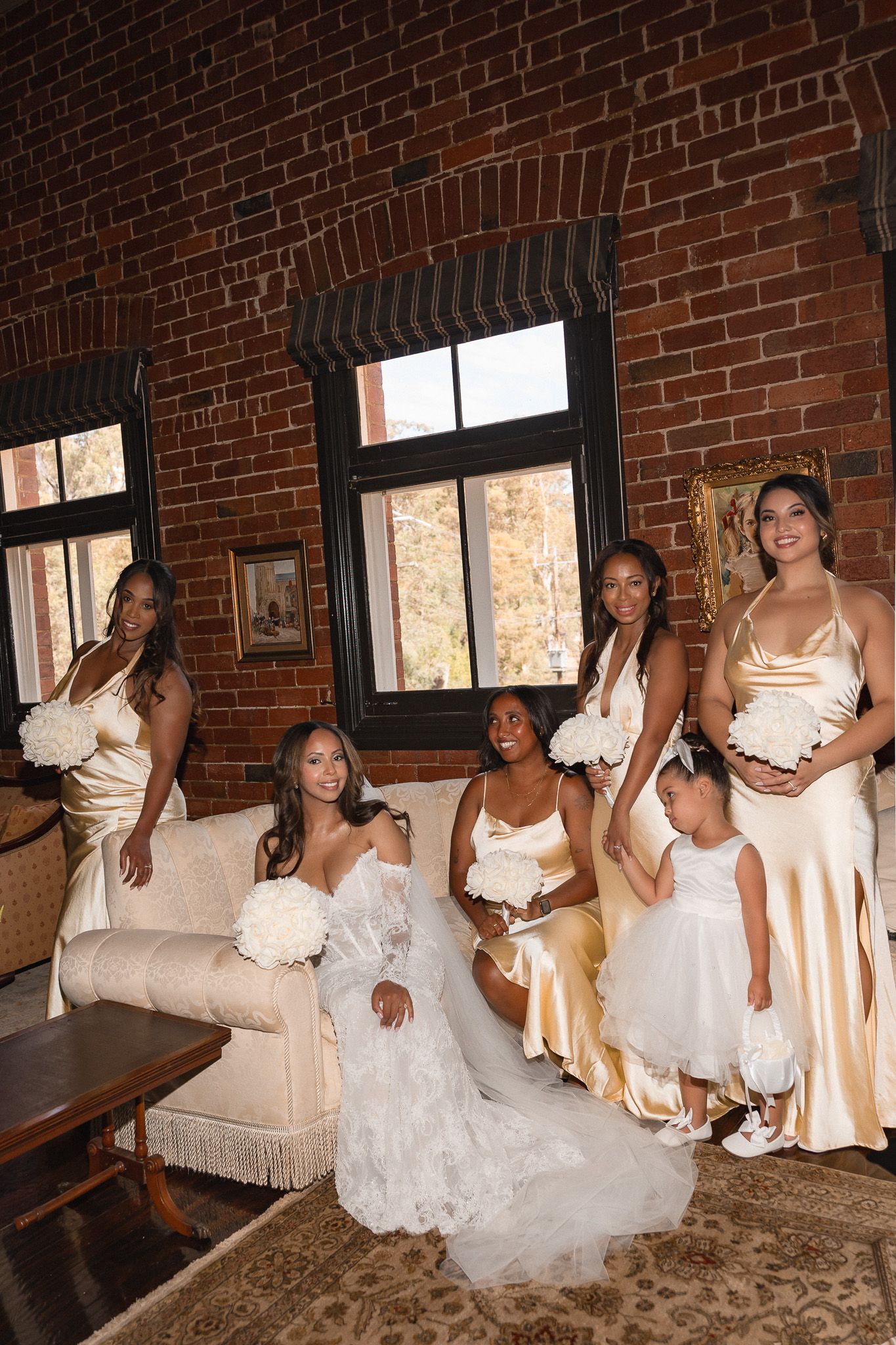 Bride in white lace gown poses with bridesmaids in gold satin dresses and flower girl in brick-wall venue.