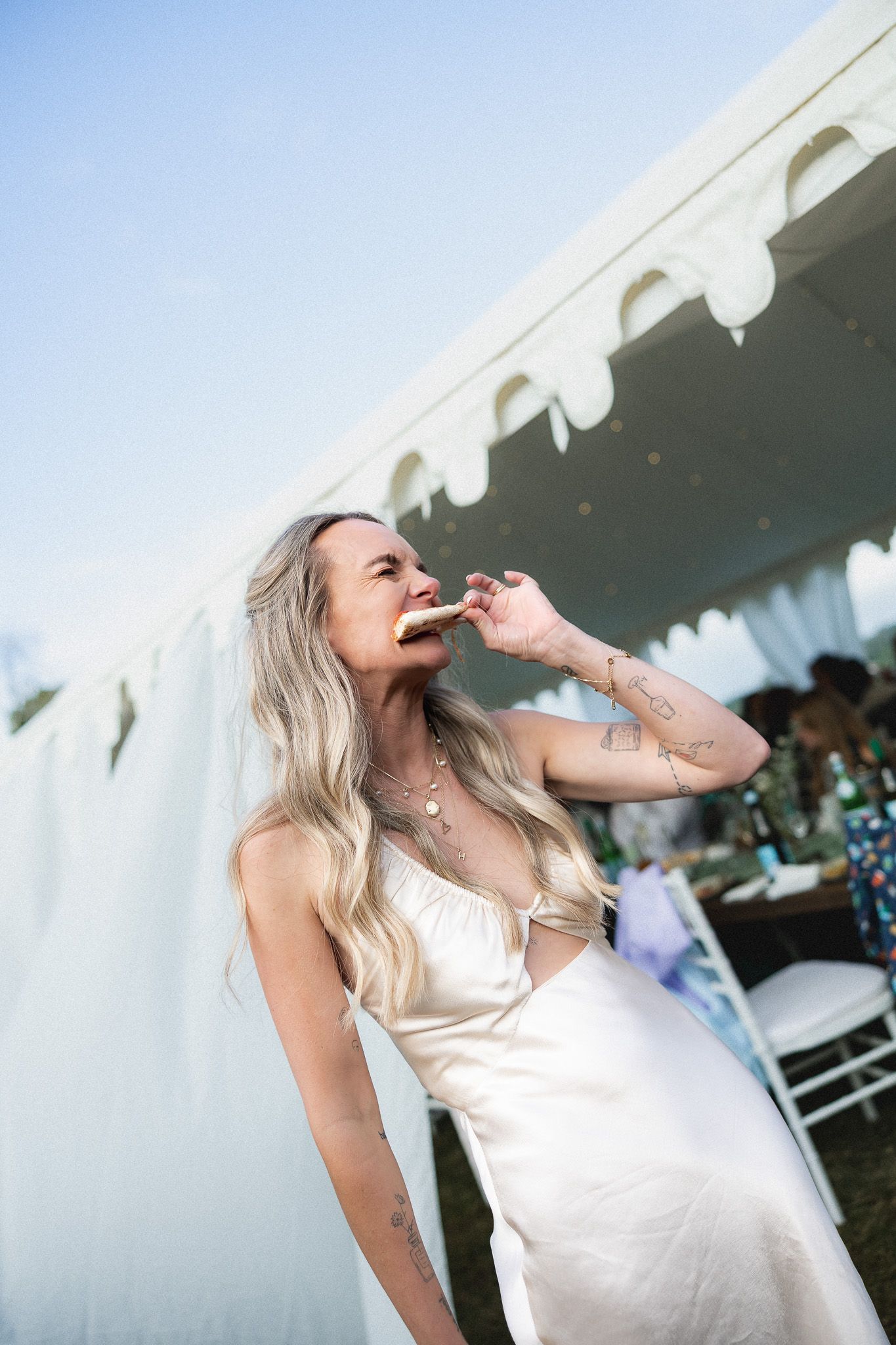 A blonde bride in a white satin dress enjoying a snack outside a wedding reception tent.