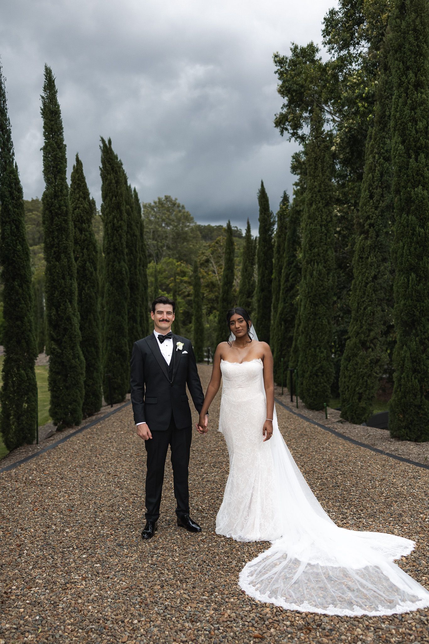Bride and groom holding hands on gravel path lined with tall cypress trees on wedding day.