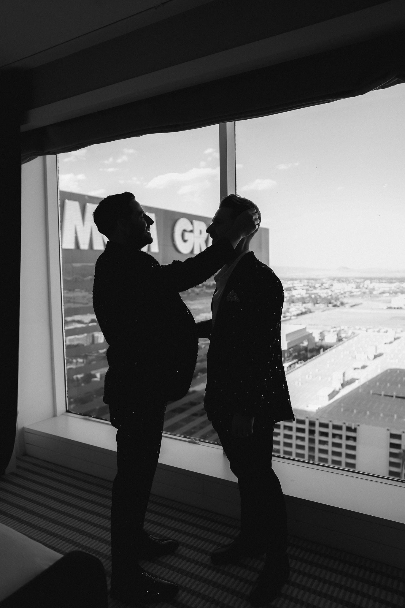 Two men silhouetted against large windows adjusting ties in a high-rise Las Vegas hotel room.