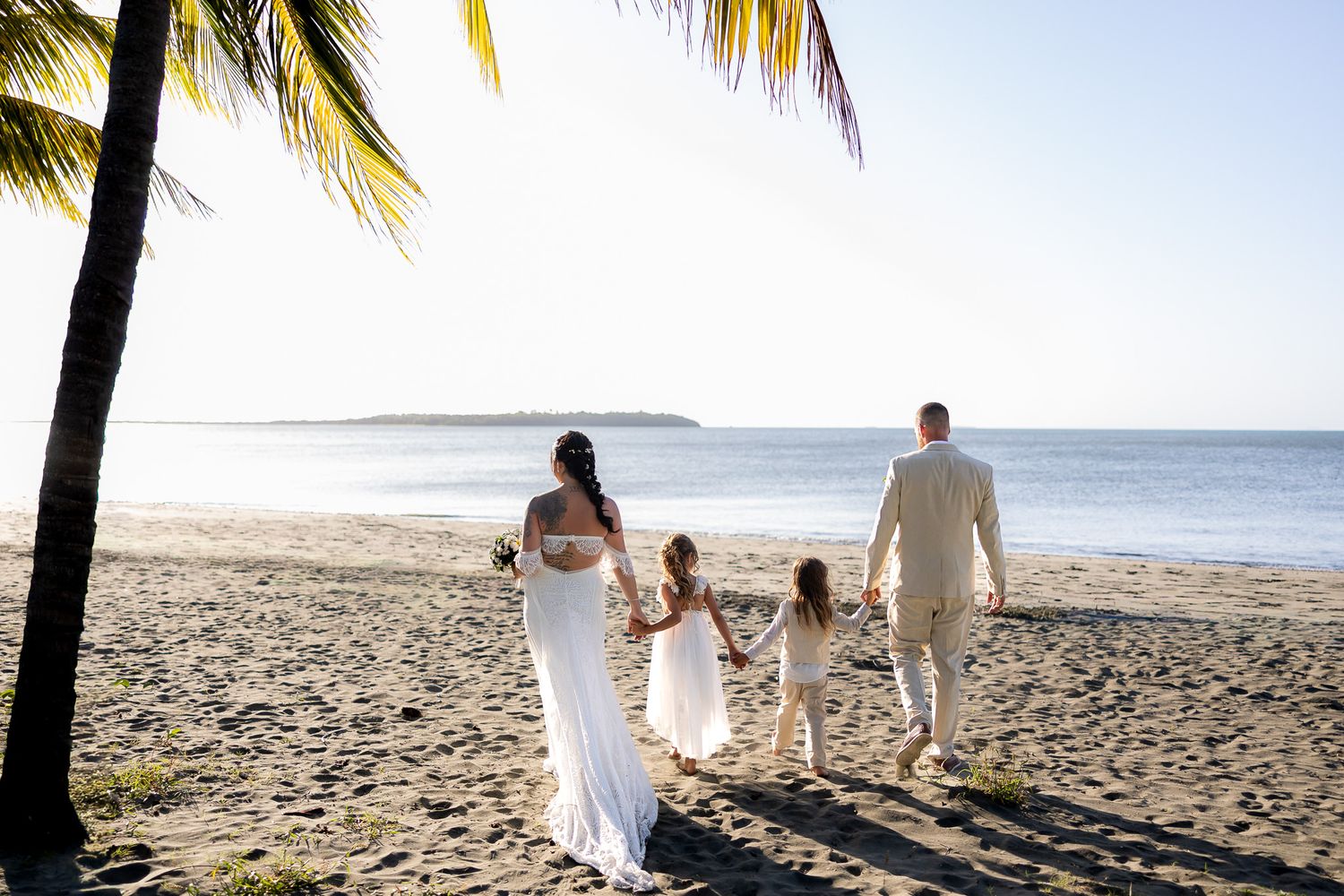 A bride and groom walk hand-in-hand with two children on a tropical beach wedding day.