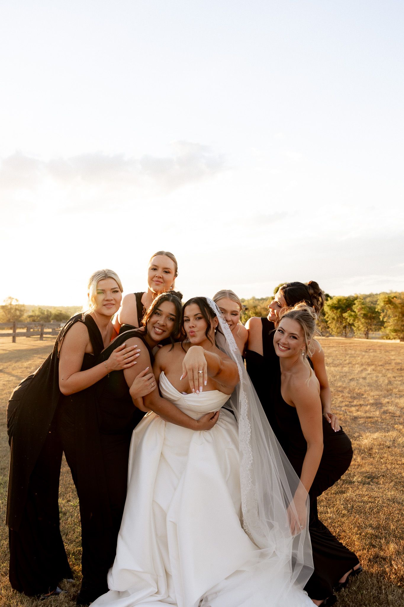 Bride in white gown poses with five bridesmaids in black dresses at golden hour in open field.