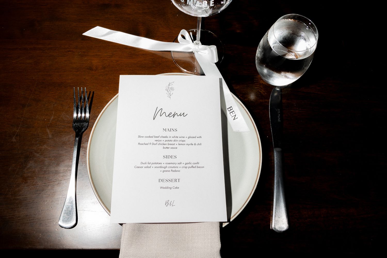 An elegant wedding place setting with a menu card, white plate, linen napkin, and glassware on a wooden table.